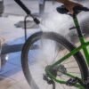 A man cleans a green bicycle with a hose, focusing on the wheel and frame outdoors.